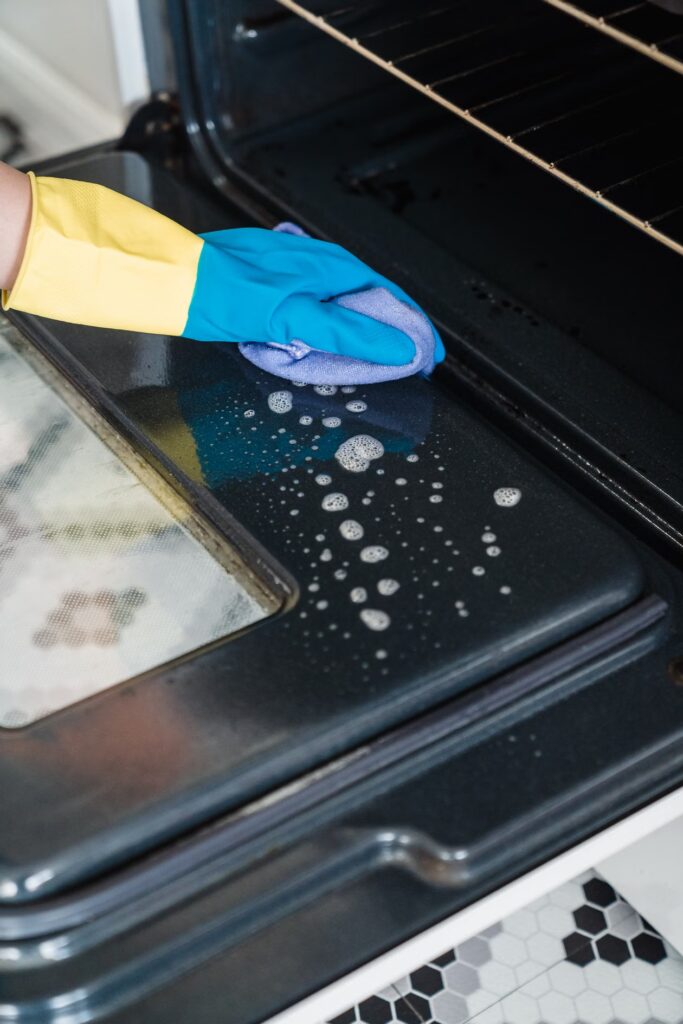Gloved hands cleaning the inside of an oven door with soapy cleaner