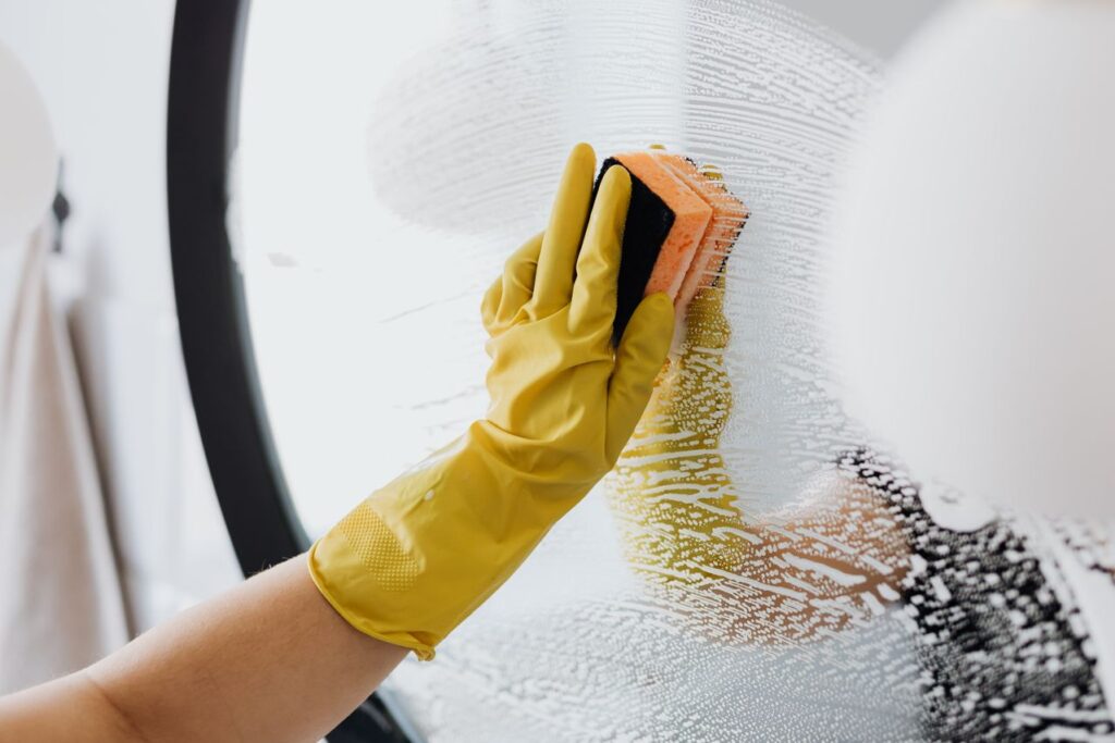 Left hand wearing a yellow rubber glove, cleaning a round mirror with a sponge and suds